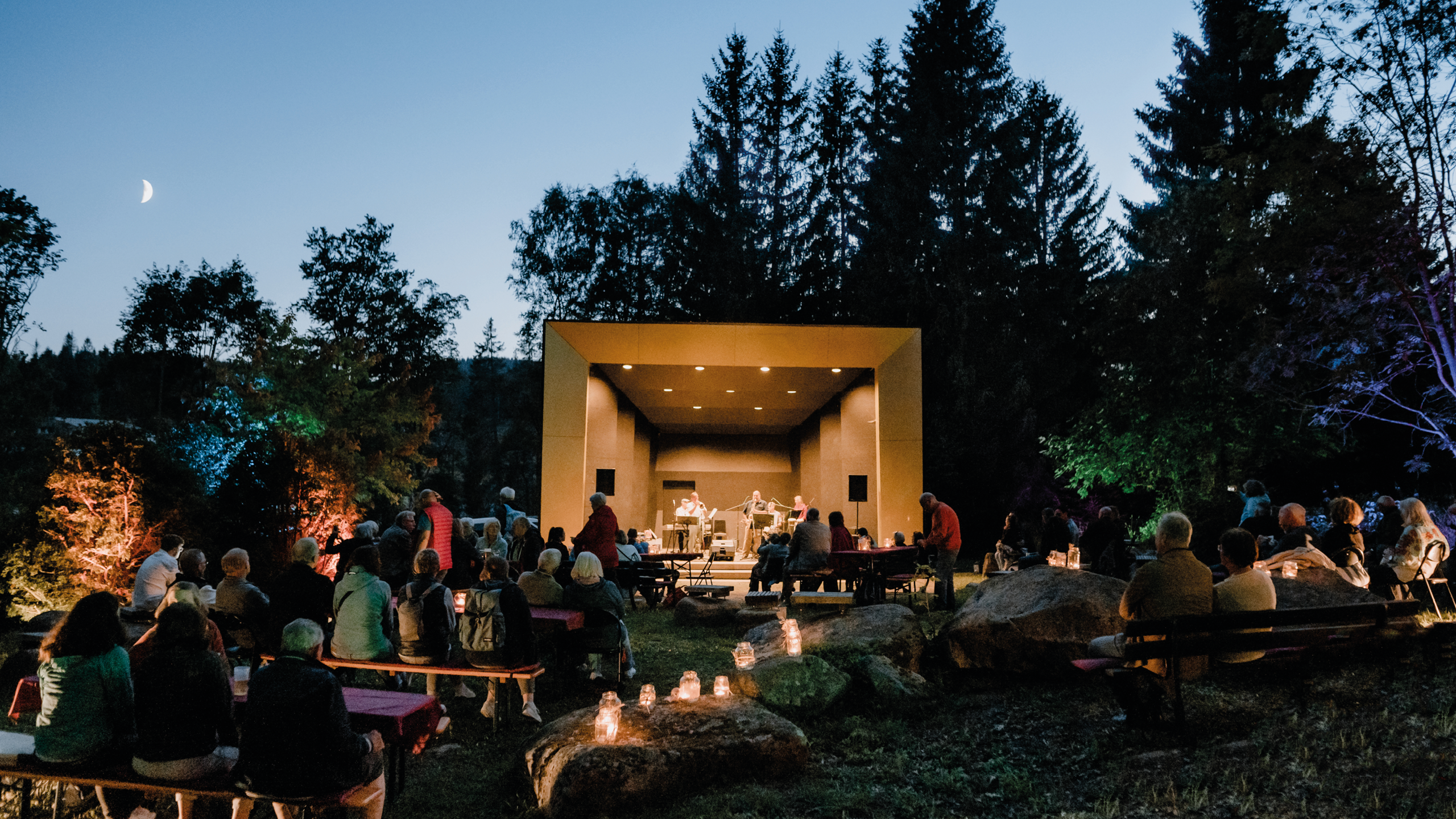 Musikpavillon im Kurpark bei Nacht. Kurpark ist mit Windlichtern geschmückt. Mond scheint am Himmel. Jazzband steht auf der Bühne. Gäste sitzen auf Bierzeltgarnituren davor. 