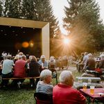 Publikum sitzt vor der Bühne des Musikpavillons im Kurpark Schierke und verfolgt die Veranstaltung zum Schierker Musiksommer. Der Pavillon erstrahlt im Gegenlicht der Sonne.